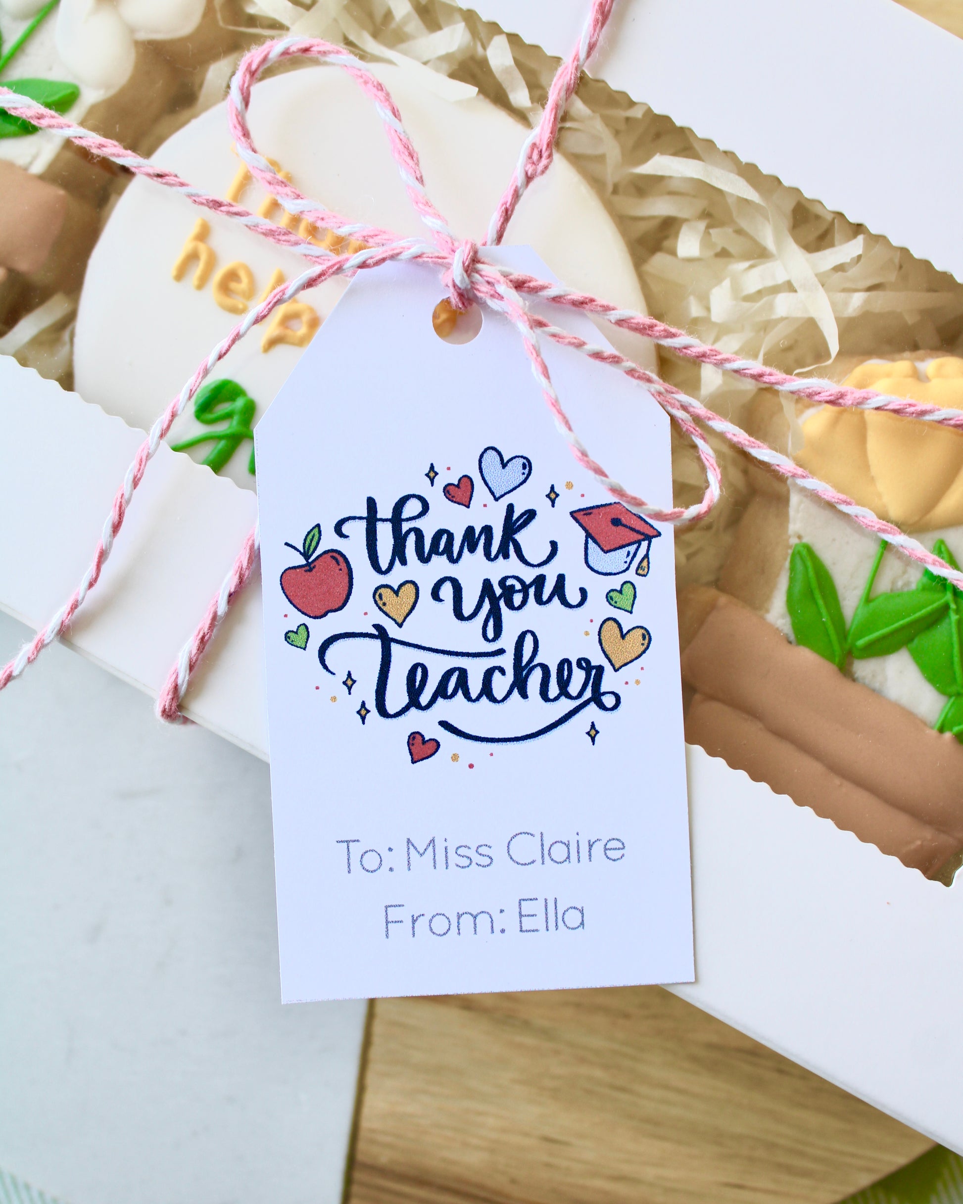 Close-up of a gift tag tied with pink and white twine to a teacher cookie gift box, reading ‘Thank You Teacher. To: Miss Claire From: Ella’ with school-themed graphics.