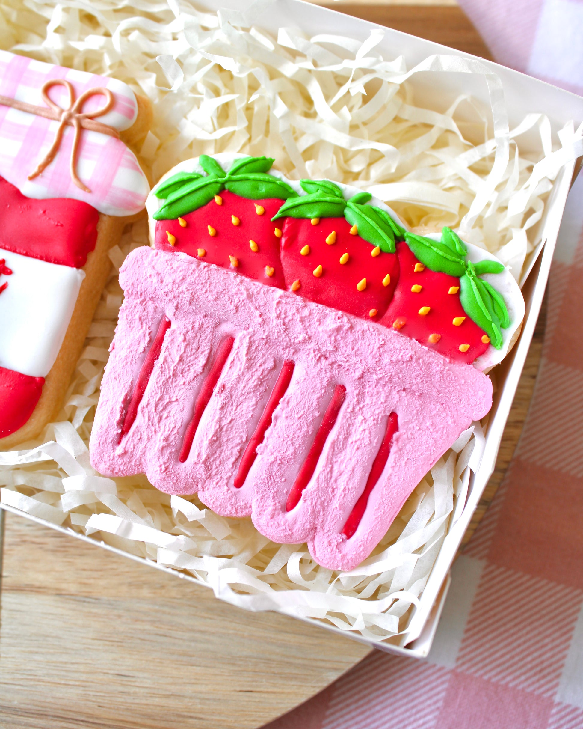 Close-up of a pink strawberry punnet cookie with textured details, decorated with three royal icing red strawberries featuring yellow seeds and green leaves.